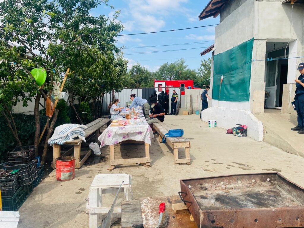 Outdoor aid station with volunteers preparing food at a long table, emergency responders in the background near a red vehicle