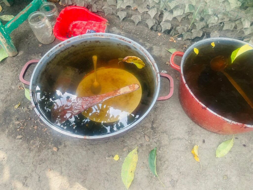 Outdoor scene with two large metal pots on dirt; left pot contains dark liquid with a yellow circular area and a wooden paddle, plus a leaf floating on top; right pot partially visible with dark liquid and a yellow spoon in it; a red plastic tub and jars sit nearby behind them.