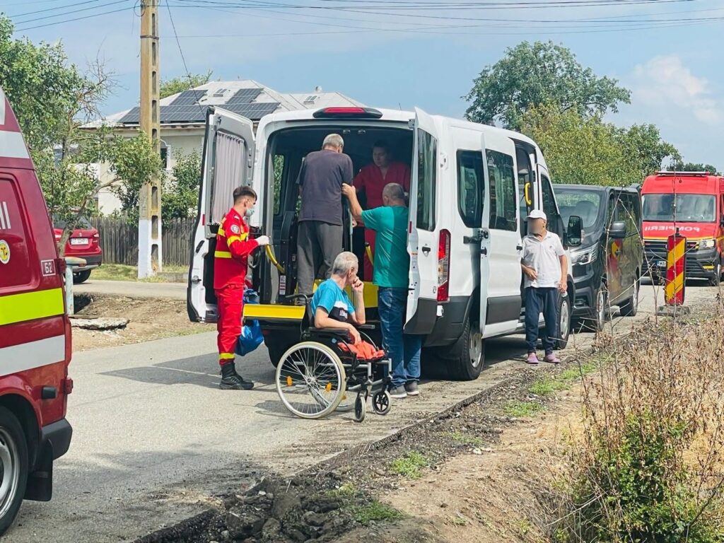Medical responders assist an elderly man in a wheelchair into a white transport van on a street, with another person helping from the doorway and a red-uniform paramedic nearby.