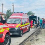 Emergency responders in red uniforms assist a person in a wheelchair beside a white van on a roadside far from a city street, with ambulances nearby
