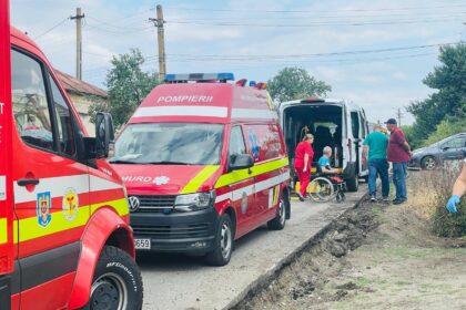 Emergency responders in red uniforms assist a person in a wheelchair beside a white van on a roadside far from a city street, with ambulances nearby