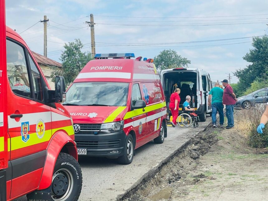 Emergency responders in red uniforms assist a person in a wheelchair beside a white van on a roadside far from a city street, with ambulances nearby