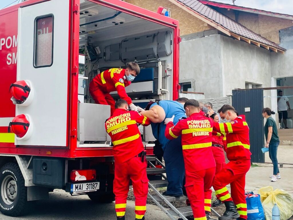 Firefighters in red uniforms assist a man into an ambulance using a stair lift, disease–smurds on scene.