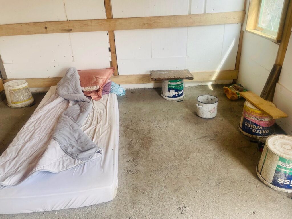 Makeshift sleeping area inside a construction shed: mattress on the floor with blankets and pillows, surrounded by paint cans and wooden planks.