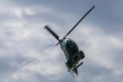 Green military-style helicopter in flight with spinning rotor blades against a cloudy sky.