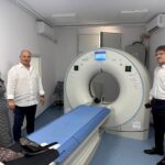 Three people stand beside a large MRI scanner in a clinical room.
