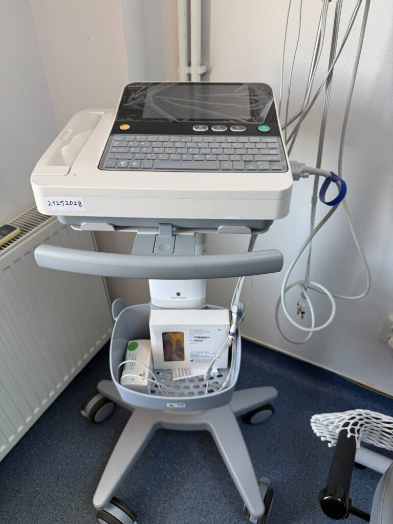Portable patient monitor on a wheeled stand in a clinical room, with a keyboard and screen under a protective cover and assorted cables nearby