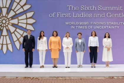 Seven world leaders stand in a line on a stage with a purple backdrop and a large emblem on the left; banner reads 'The Sixth Summit of First Ladies and Gentlemen'.