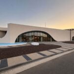 Modern white building with a large curved glass entrance and water feature in front at sunset