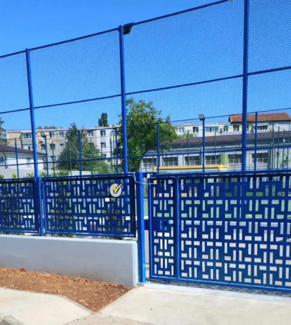 99882-99882-36 - Jurnalul Olteniei – Știri din Craiova și Oltenia Blue perforated metal fence and gate enclosing a sports court, set against a clear blue sky.