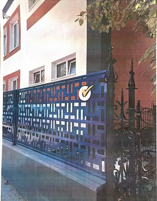99883-99883-36 - Jurnalul Olteniei – Știri din Craiova și Oltenia Blue decorative metal gate with a geometric cutout pattern beside a pale building, featuring a circular yellow-and-white checkmark sticker.