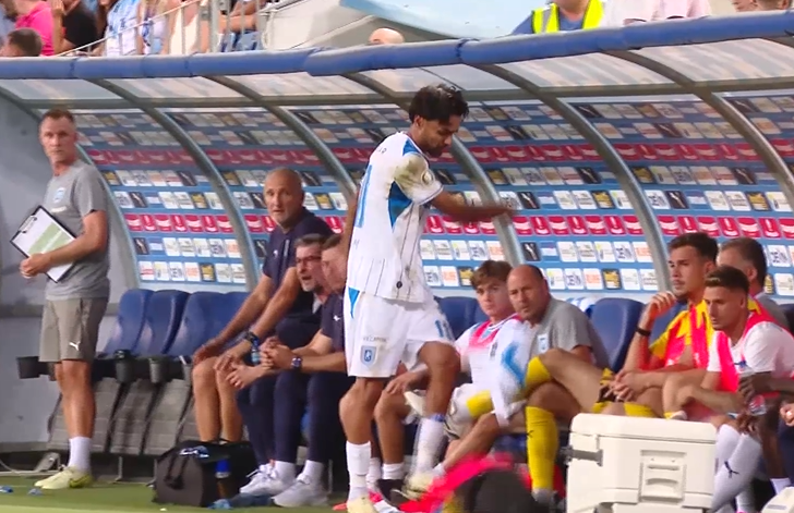 Captură de ecran 2026-08-14 084713 - Jurnalul Olteniei – Știri din Craiova și Oltenia Soccer player in white and blue standing near team bench, others seated and watching on the sideline.