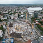 Aerial view of a circular construction site with several cranes in a city neighborhood and a large white stadium in the background.