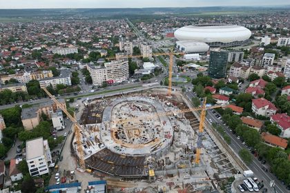 Aerial view of a circular construction site with several cranes in a city neighborhood and a large white stadium in the background.
