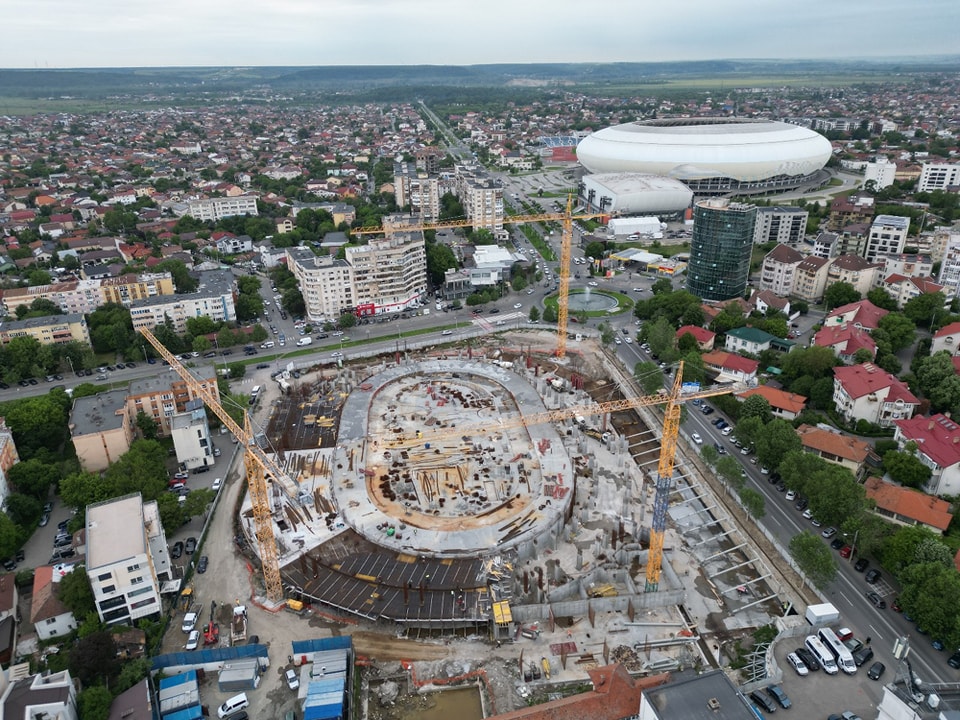 Aerial view of a circular construction site with several cranes in a city neighborhood and a large white stadium in the background.