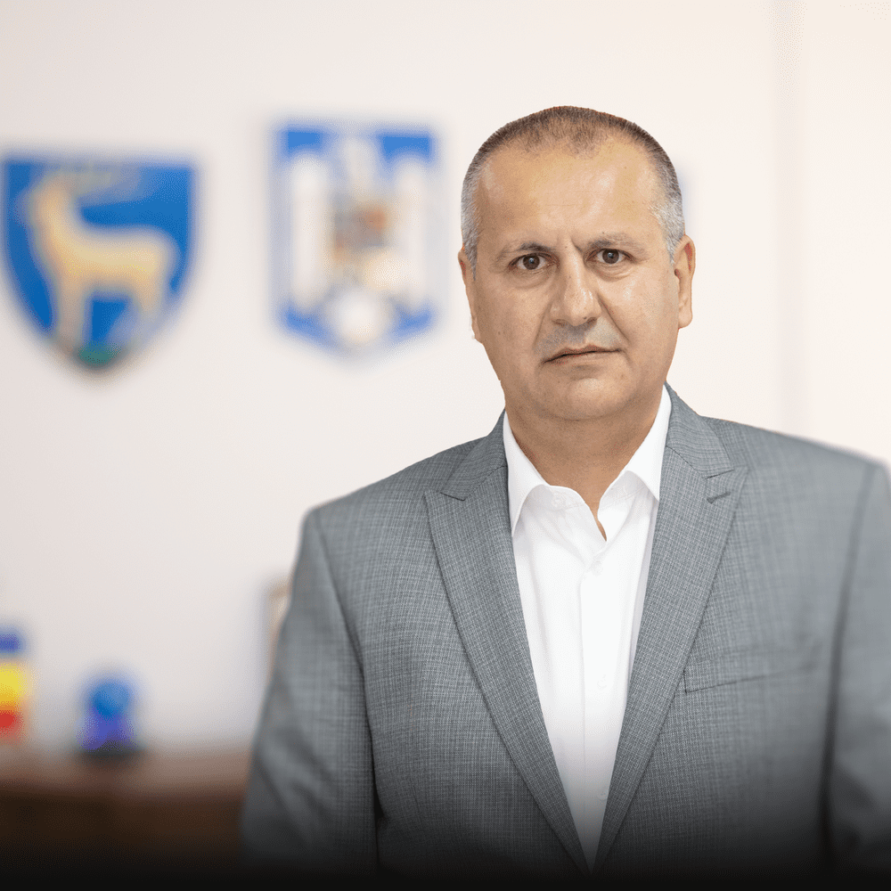 Professional man in a gray suit posing in an office with blurred blue emblems on the wall behind him.