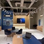 Optical store interior with blue walls, wooden display units, and shelves of eyeglasses around a reception desk area.
