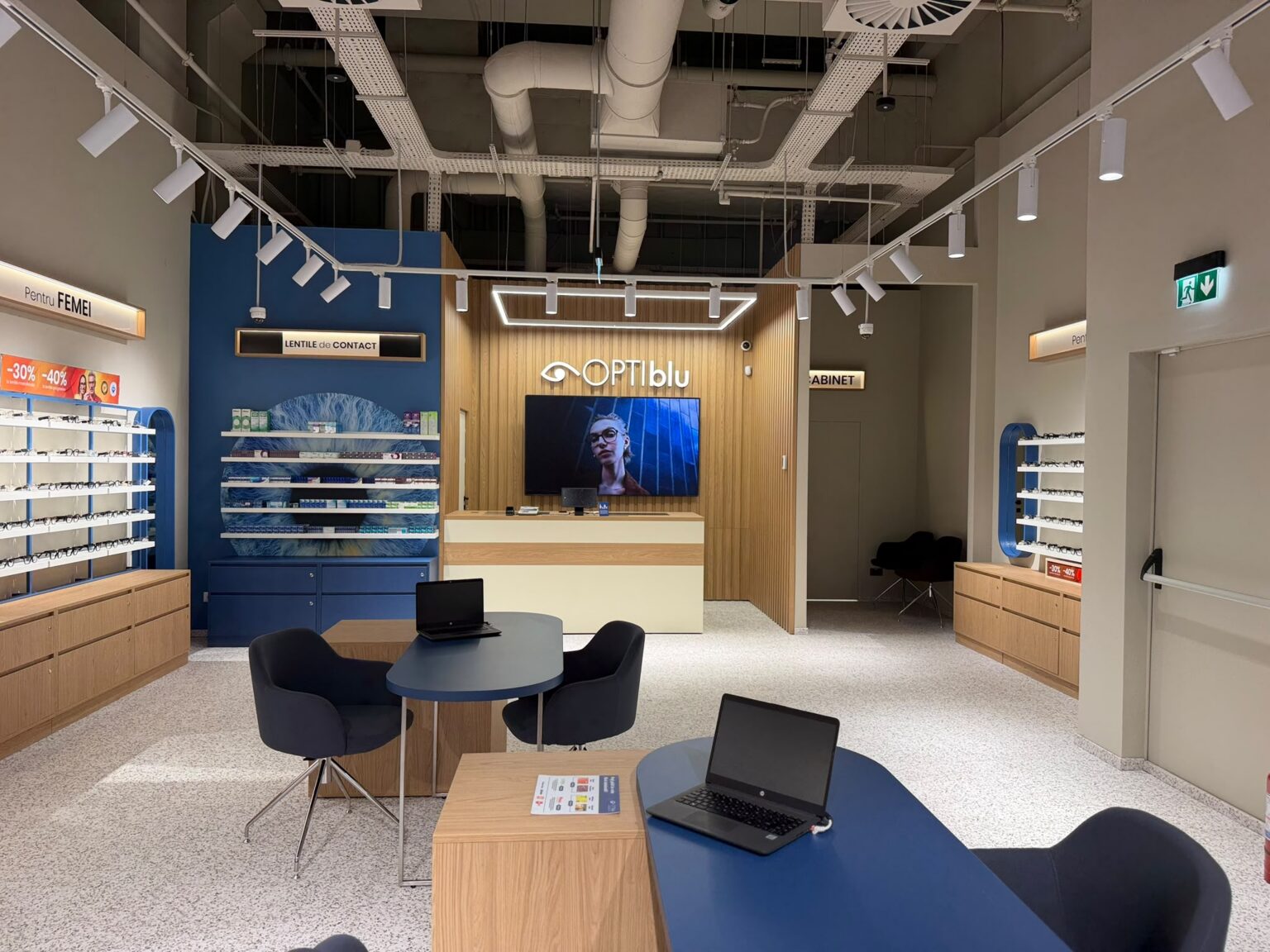 Optical store interior with blue walls, wooden display units, and shelves of eyeglasses around a reception desk area.
