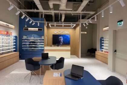 Optical store interior with blue walls, wooden display units, and shelves of eyeglasses around a reception desk area.