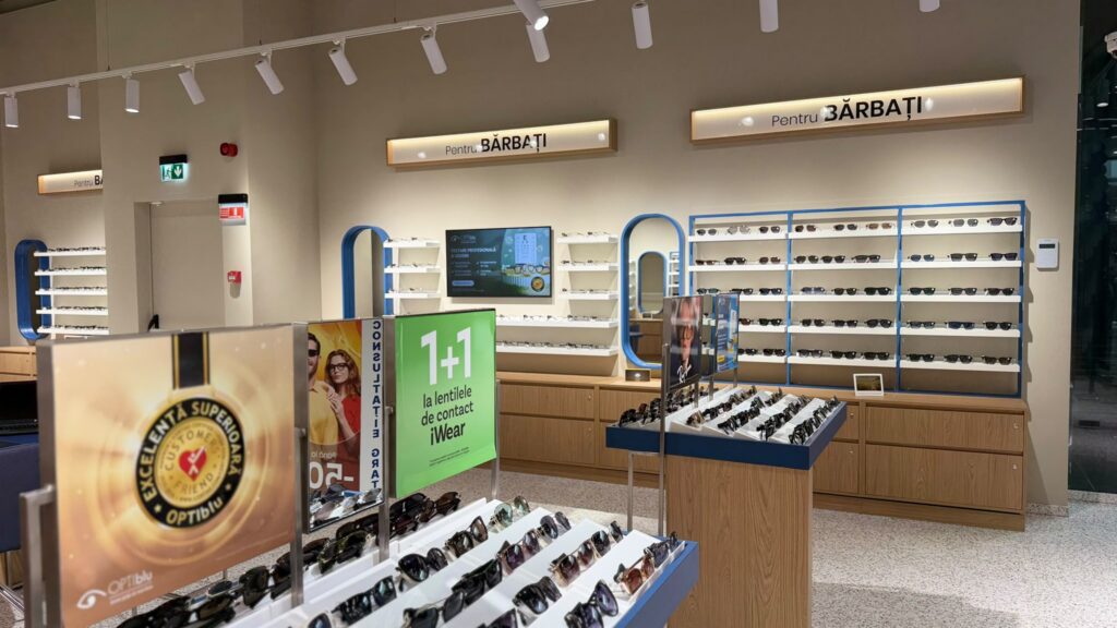 Eyewear store interior with rows of sunglasses on blue-and-wood displays; signs say "Pentru BĂRBAȚI" (For Men).
