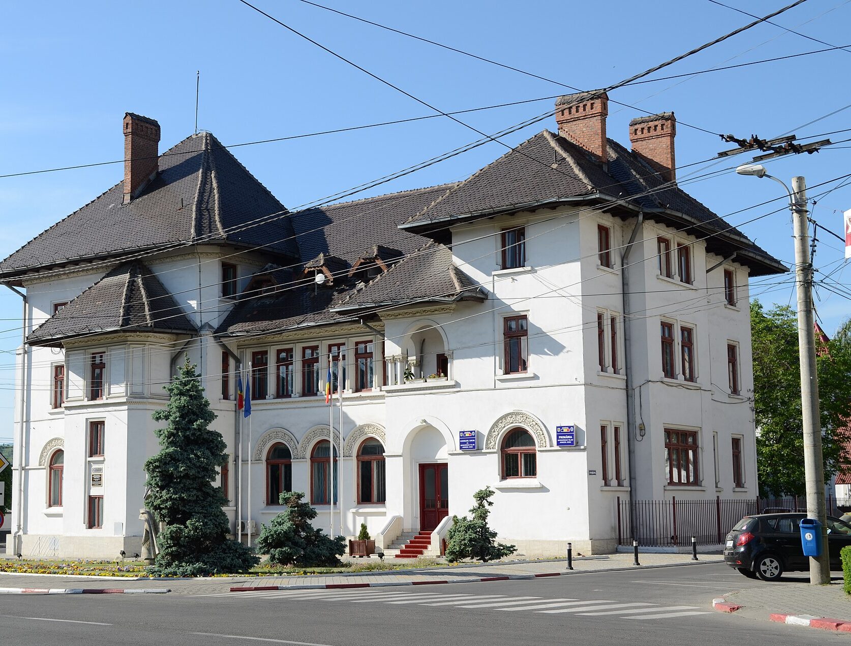 White multi‑story building with a steep, dark tiled roof and brick chimneys, on a sunny street with overhead power lines.