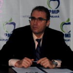 Man in a dark suit and glasses sits at a desk with a pen, in front of a backdrop with PMP logos.