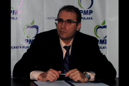 Man in a dark suit and glasses sits at a desk with a pen, in front of a backdrop with PMP logos.