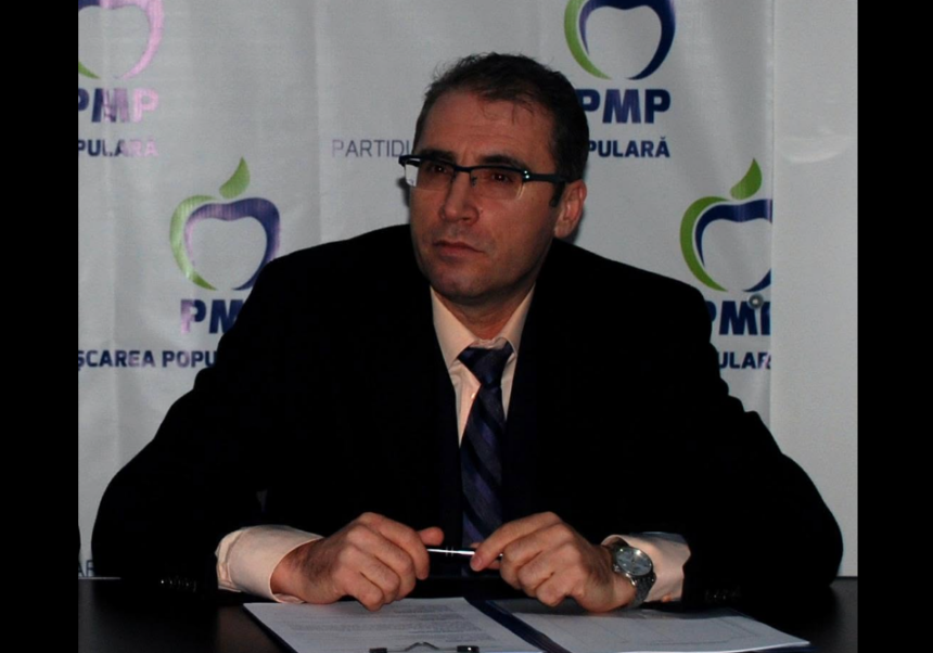 Man in a dark suit and glasses sits at a desk with a pen, in front of a backdrop with PMP logos.