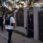 Romanian police officer in a vest walking toward a gated residential entrance beside a white van in a gated neighborhood