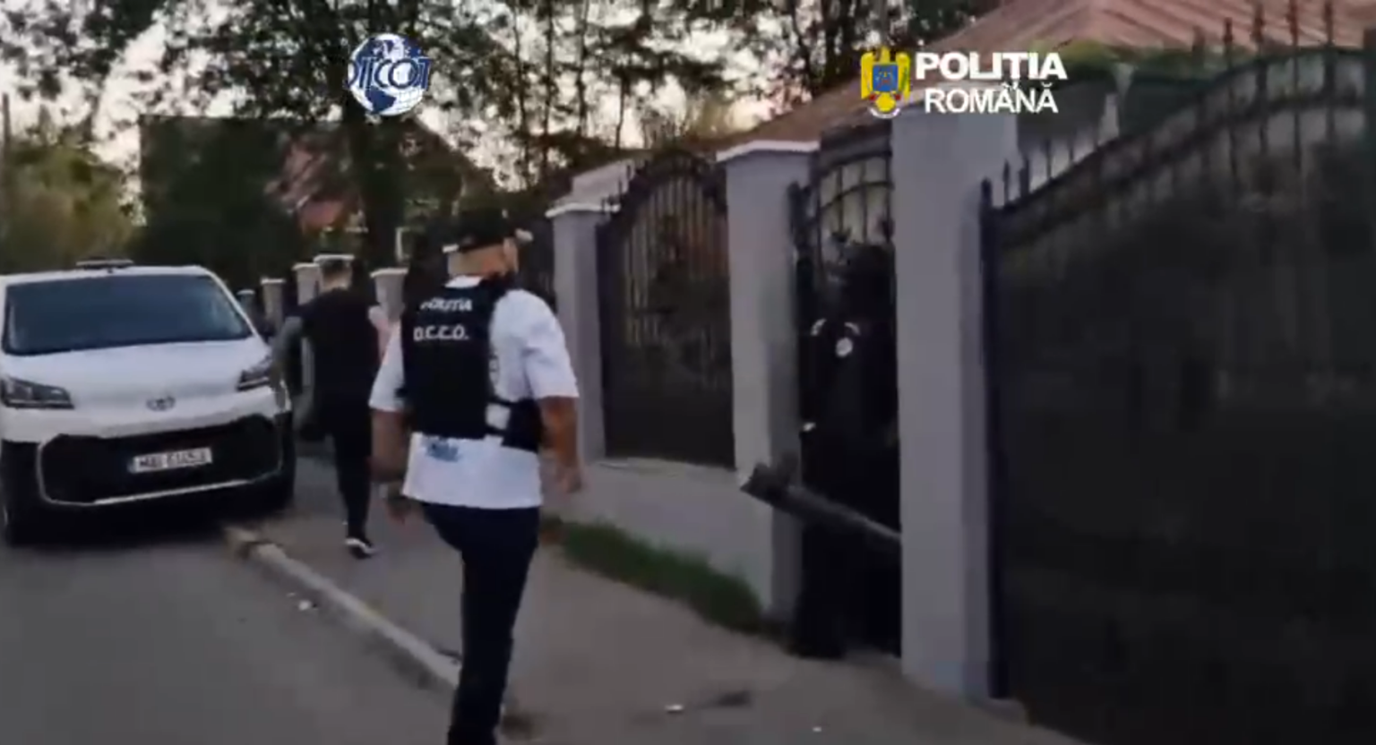 Romanian police officer in a vest walking toward a gated residential entrance beside a white van in a gated neighborhood