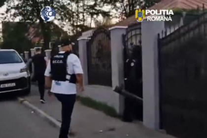 Romanian police officer in a vest walking toward a gated residential entrance beside a white van in a gated neighborhood