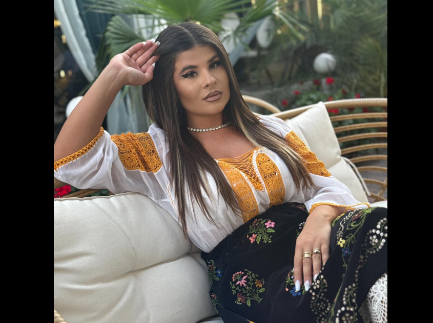 Woman reclining on a cream cushion wearing a white embroidered blouse in orange and a dark floral skirt, posing outdoors with palm plants in the background.