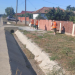 Three road workers in orange safety vests stand along a sidewalk beside a red wooden fence, near a concrete drainage ditch.