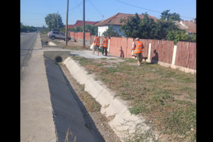 Three road workers in orange safety vests stand along a sidewalk beside a red wooden fence, near a concrete drainage ditch.