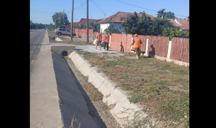 Three road workers in orange safety vests stand along a sidewalk beside a red wooden fence, near a concrete drainage ditch.