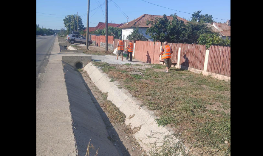 Three road workers in orange safety vests stand along a sidewalk beside a red wooden fence, near a concrete drainage ditch.