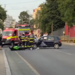 Emergency responders and a red ambulance attend a car crash on a city street; a dark car is involved, bystanders nearby.