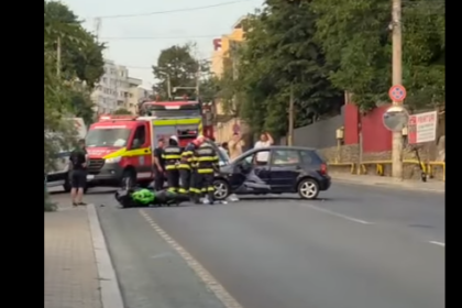 Emergency responders and a red ambulance attend a car crash on a city street; a dark car is involved, bystanders nearby.
