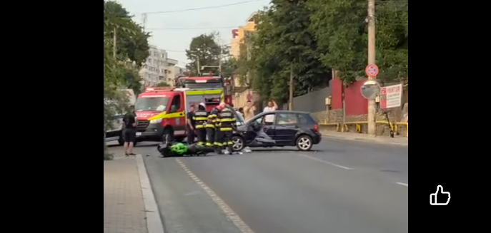 Emergency responders and a red ambulance attend a car crash on a city street; a dark car is involved, bystanders nearby.