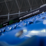 Blue stadium seating with a black banner reading 'UEFA CONFERENCE LEAGUE' along the stands inside a stadium.