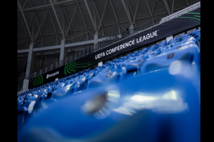 Blue stadium seating with a black banner reading 'UEFA CONFERENCE LEAGUE' along the stands inside a stadium.