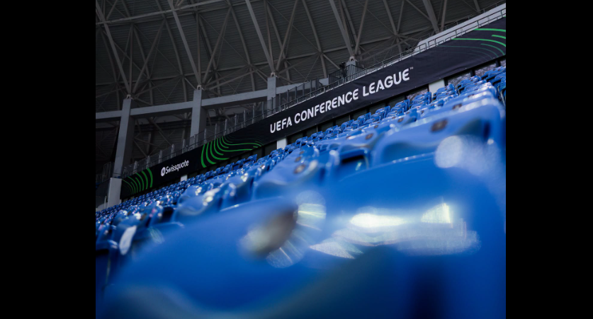 Blue stadium seating with a black banner reading 'UEFA CONFERENCE LEAGUE' along the stands inside a stadium.