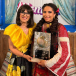 Two women pose with a glass award featuring a shell, smiling in front of a colorful backdrop.