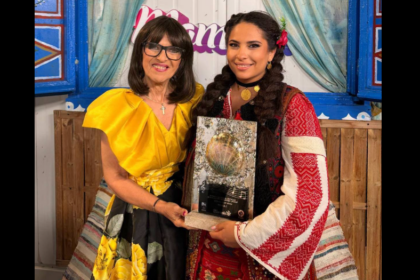 Two women pose with a glass award featuring a shell, smiling in front of a colorful backdrop.