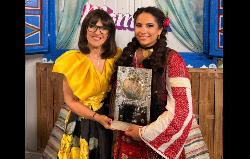 Two women pose with a glass award featuring a shell, smiling in front of a colorful backdrop.