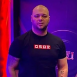 Man with a shaved head in a black DSP2 shirt standing in a purple-lit studio backdrop.