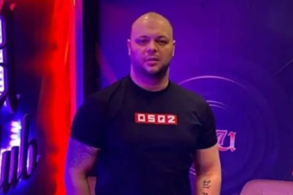 Man with a shaved head in a black DSP2 shirt standing in a purple-lit studio backdrop.