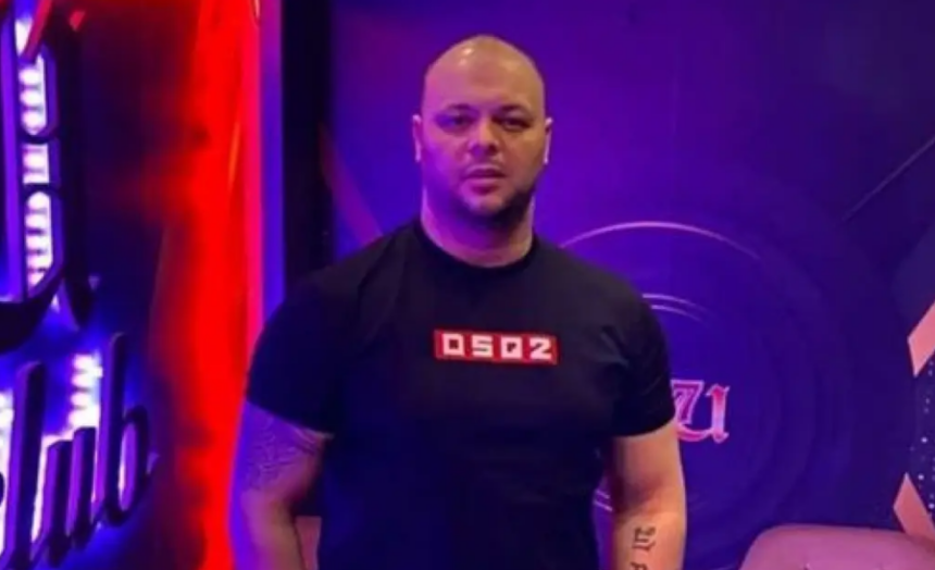 Man with a shaved head in a black DSP2 shirt standing in a purple-lit studio backdrop.