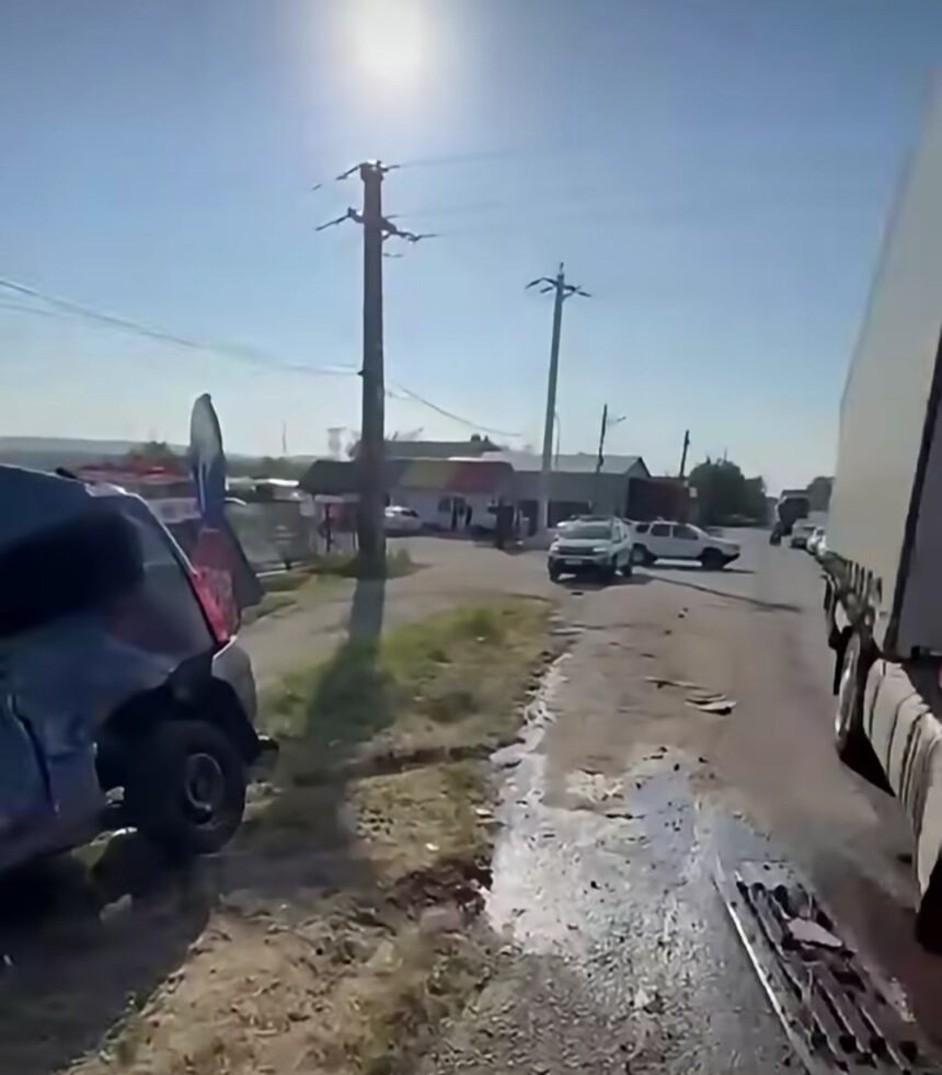 Sunlit urban street with a large truck on the right, a white SUV ahead, and a blue van parked on the left amid muddy patches.