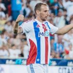 Soccer player in white jersey with blue and red stripes celebrates with arms raised as crowd cheers behind him
